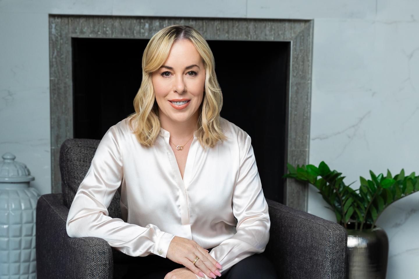 CEO Amy McKenna wearing a white blouse with her hands crossed, sitting in a grey armchair and smiling at the camera